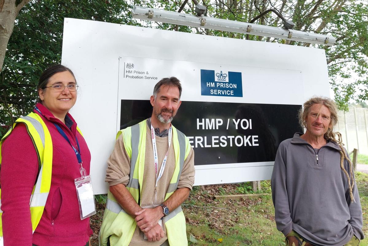 Eve, Lee and Steve are smiling, standing in front a white sign which says HMP Erlestoke 