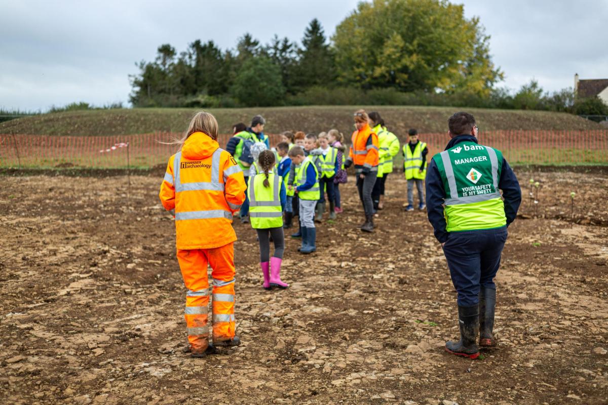 A Wessex Archaeology staff member in orange hi-vis talks to a group of school children