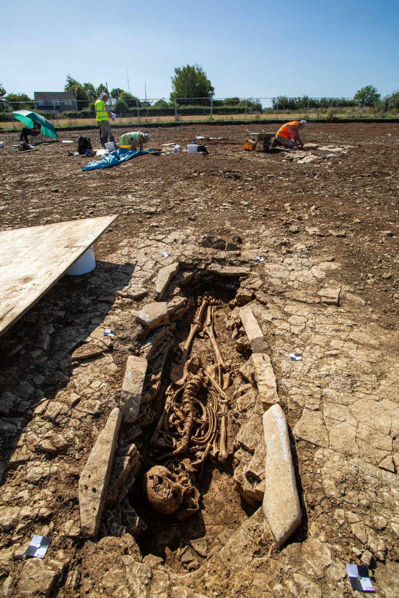 Photograph of archaeologists working on site. A grave containing a skeleton has been uncovered in the foreground