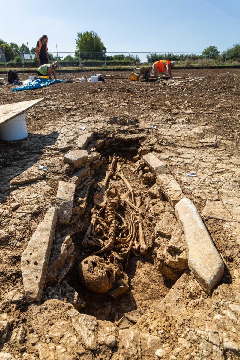 A skeleton is seen within a grave cut, with archaeologists working in the background