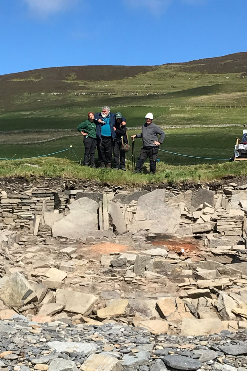 The site team at the Orkney archaeological site stand on a grassy ledge overlooking the site whilst waiting for the right weather conditions to take a photo